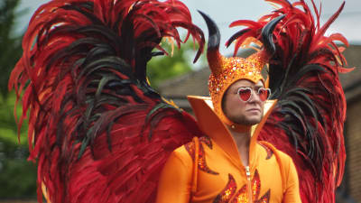 Elton John (Taron Egerton) iklädd en paljettbeströdd orangedräkt med stora röda vingar och en mössa med paljetter och horn.