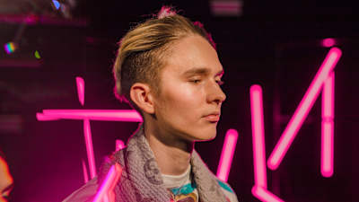 Vaaleahiuksinen nuori mies katsoo alaviistoon vaaleanpunaisten neontankojen edessä, kuva rajattu hartioista alaspäin.
