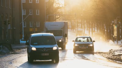 Tre bilar kör på Backasgatan i Helsingfors. Solen glänser på vägytorna.