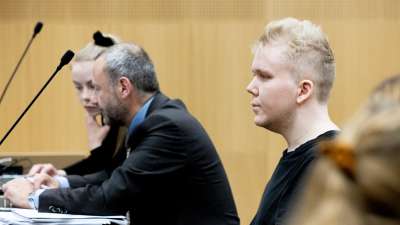 Aleksanteri Kivimäki fotograferad från sidan medan han sitter vid ett svart bord i en rättssal. Bredvid honom sitter en man och en kvinna.