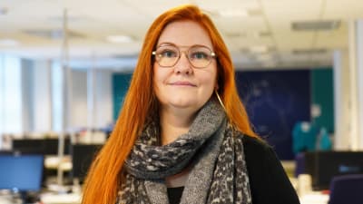 En kvinna med långt rött hår och glasögon tittar in i kameran. I bakgrunden syns ett tomt kontorslandskap.