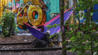 Man ligger i färggrann hängmatta fäst mellan ett par träd som sticker upp mellan järnvägsspår. I bakgrunden färggann graffittivägg.