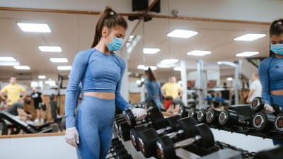 En kvinna i munskydd lyfter upp vikter på ett gym.