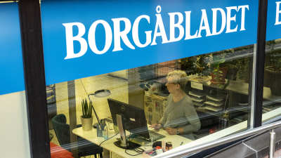 Ett fönster med texten Borgåbladet. Bakom rutan jobbar en kvinna vid dator.