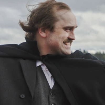 Pukuun ja viittaan pukeutunut mies pitää kädessään mustaa, isolieristä hattua ja heristää nyrkkiä irvistäen. Taustalla järvi, ranta siintää taka-alalla.