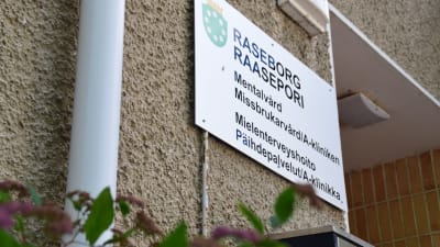 Skylt där det står Raseborg och stads vapen. Under den står det mentalvård, missbrukarbvård/a-kliniken. Skylten sitter på ett hus brevid ingången.