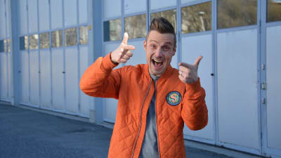 Programledaren Jontti Granbacka står på en parkering iklädd en orange jacka med logon för nya barnprogrammet Superhjälparna.