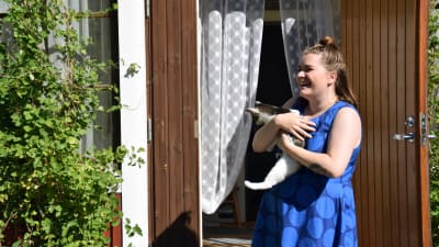 En kvinna i blå klänning står med en katt i famnen. I bakgrunden skymtar en ingång till ett hus.