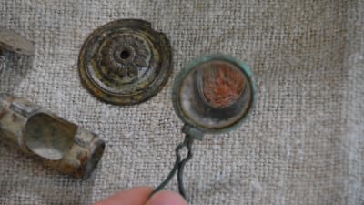 På bilden används ett gammalt förstoringsglas hittat i åkern för att förstora en läppstiftsburk. Bredvid ligger ett mantelspänne och en del av en tvärflöjt.