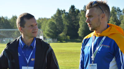 Johan Byholm och Putte Lindroos stpr bredvid varandra och talar på en fotbollsplan. De bär båda fotbollskläder.