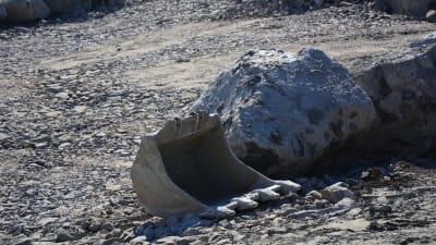 En grävskopa ligger på marken bredvid ett stort stenblock.
