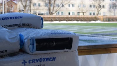 Plastsäckar i förgrunden, i bakgrunden syns en grön spelplan med lite snö. Solen skiner på planen. Längs borta i bakgrunden ett flervåningshus.