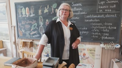 Dam i vit skjorta, svart kofta och glasögon står bakom en kateder, i bakgrunden svart tavla med teckningar och texter skrivna med krita.