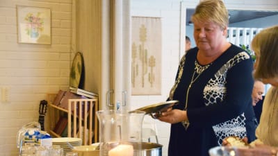 En kvinna står med ett kastrulllock i handen vid en bord där det serveras mat. Brevid henne står en annan kvinna och tar åt sig mat. Kvinnan på bilden heter Mona Andersson.