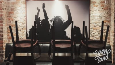 Restaurangbord med uppstaplade stolar, i bakgrunden ett fotografi av människor som dansar med händerna i luften.