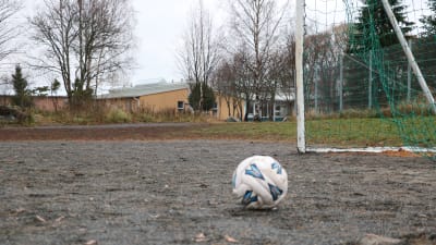 En fotboll ligger på en grusplan. I bakgrunden skymtar en skolbyggnad.