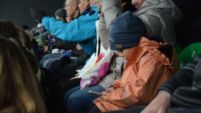 En pojke sitter och tittar på sin telefon på en läktare där alla vuxna följer med matchen.