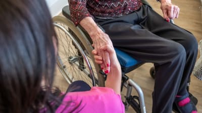 En vårdare håller i en äldre persons hand. Den äldre sitter i rullstol.