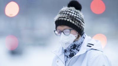 En person i munskydd går ute i vintervädret. Hon har immiga glasögon på sig och mössa.