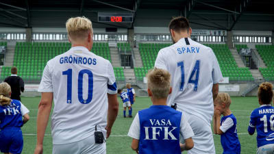 Vi ser Janne Grönroos och Tim Sparv bakifrån i blå-vita fotbollsskjortor med efternamnen på ryggen. Några fotbollsjuniorer i Vasa IFK-spelarskjortor syns i bildens nedre kant.
