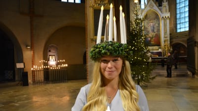 Åbos lucia 2020 och 2021 Janina Karrento, en dam med långt ljust hår och en ljuskrona på huvudet, tittar in i kameran. Hon står i Åbo Domkyrka med en stor julgran i bakgrunden.