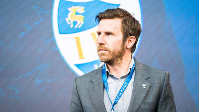 Daniel Norrmén i grå kostym med IFK Mariehamns logo bakom sig.