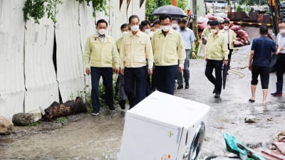 Premiärminister Han Duck-soo leder en grupp som inspekterar översvämningsskador på en gata där vatten rinner ner och med bråte - inklusive en tvättmaskin - har samlats. I bakgrunden skymtar högar av sandsäckar.