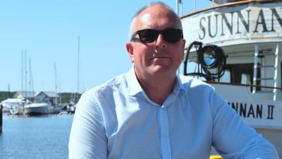 Företagare Tom Nylund framifrån sittande på en brygga vid restaurangbåten Sunnan en solig sommardag. Hav i bakgrunden.