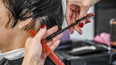 Närbild på två händer som håller på att klippa en person med mörkt kort hår.