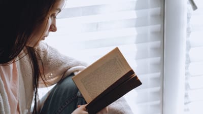 Ung kvinna sitter vid ett fönster och läser en bok