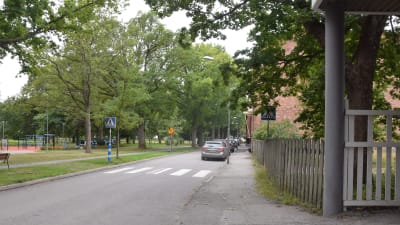 En gata vid ett övergångsställe. På ena sidan en motionspark, på andra sidan ett rött tegelhus, staket och några bilar som står parkerade.