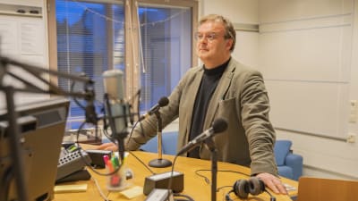 En man i glasögon och ljus kavaj står vid ett bord i en radiostudio. Han ser allvarlig ut. På bordet mikrofoner och en datorskärm.