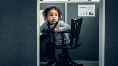 Ett litet barn tittar fram bakom en skärm i ett anonymt kontorslandskap.