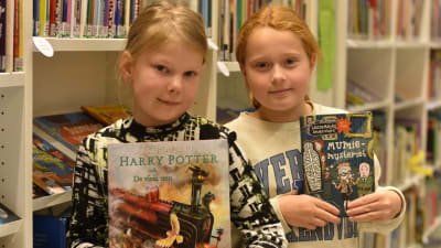 Två flickor står vid en hylla i ett bibliotek och håller i varsin favoritbok.