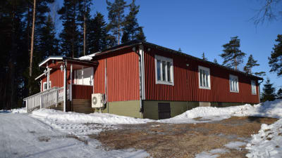 En bild på ett rött hus som står på en kulle. Det röda huset är i ett plan och är omgivet av skog. Huset är från 60-talet.
