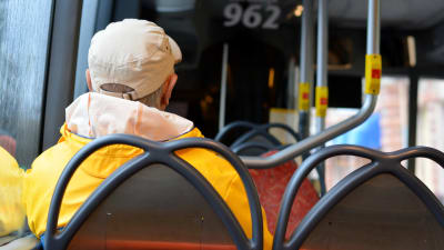 En man i gul regnrock som sitter i en buss med ryggen vänd mot kameran. 