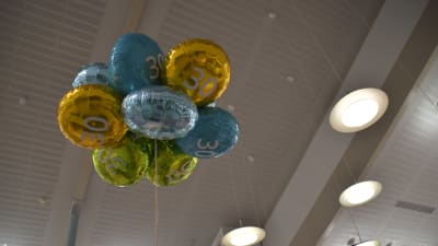 Folieballonger fyllda med helium i en hopsamlad "bukett" svävar mot ett vitt trätak.  Ballongerna är i gult, turkos och grönt. De har siffran 30 i mitten.