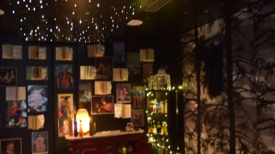 Ett mörkt rum med böcker och affischer på väggarna. Stämningsbelysning i form av ljusgirlanger. Bibliotek, sagorum.