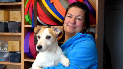 En kvinna står med en liten, ljus hund i famnen, i bakgrunden syns rullar med remmar i olika färger.