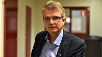 Man med glasögon och blå kragskjorta sitter på en stol och ser in i kameran.