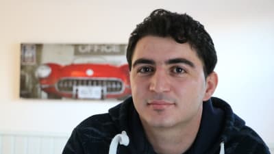 En syrisk ung man med mörkt hår. I bakgrunden skymtar en tavla med en röd amerikansk bil som motiv.