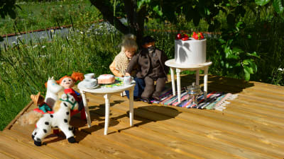 En Pippi-docka, ett Mumintroll-gosedjur och två dockor är placerade på ett brunnslock ute i en trädgård.