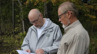 Två män står framför ett skogsparti och tittar på ett papper.