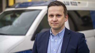 En man med polisbricka runt halsen står utomhus med en polisbil i bakgrunden.