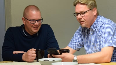 Två män dricker kaffe vid ett bord och kollar på en smartphone och ler.