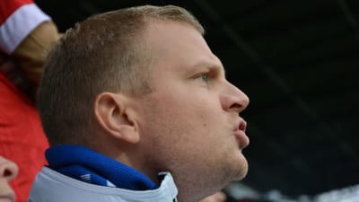 En man med blå halsduk och ljusblå jacka hejar på sitt lag på en fotbollsmatch. Läpparna formas till ordet "Skjut!".