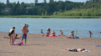 Många människor ligger på en sandstrand och solar sig. Några barn leker i vattnet.