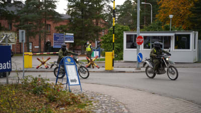 Två mopedister i militäruniform åker ut förbi en öppen bom där en person i uniform står och bevakar vid ett vaktbås.
