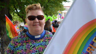 En person med färggrann skjorta och prästkrage håller i en banderoll med regnbågsfäger.