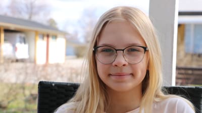 En elvaårig flicka med långt blont hår och glasögon sitter ute på en stol.